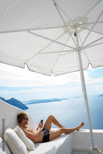 Man relaxing with his mobile phone on a white balcony with a dramatic scenic view of the Mediterranean Sea and Santorini caldera © PeskyMonkey