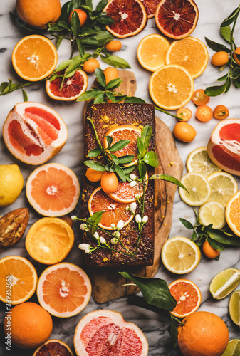 Citrus Mediterranean loaf cake. Flat-lay of almond pound cake with fresh oranges, lemon, kumquat, grapefruit and blossom flowers on board over grey marble background, top view. Vegan dessert concept