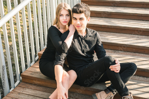 Beautiful young multiracial couple, student couple in love, sit wooden staircase in the city. Beautiful Turkish brunette guy hug a Caucasian girl in black clothing. Date young people in sunny weather © Elizaveta