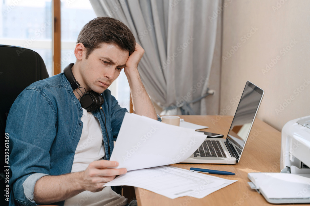 Thoughtful young man sitting at table with laptop and looking seriously at papers. Lost job due to crisis or coronavirus quarantine, thinking about how to pay bills. Remote work and distance education