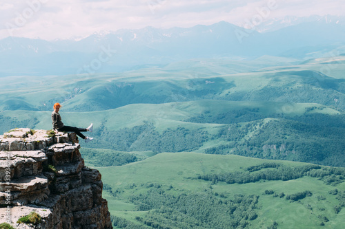 A young woman sits on the edge of a cliff against the backdrop of the Caucasus mountains and dangles her legs