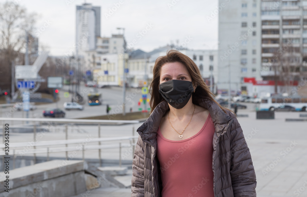 Woman of European appearance in a black medical mask on her face from the coronavirus looks at the background of empty Grod and stre