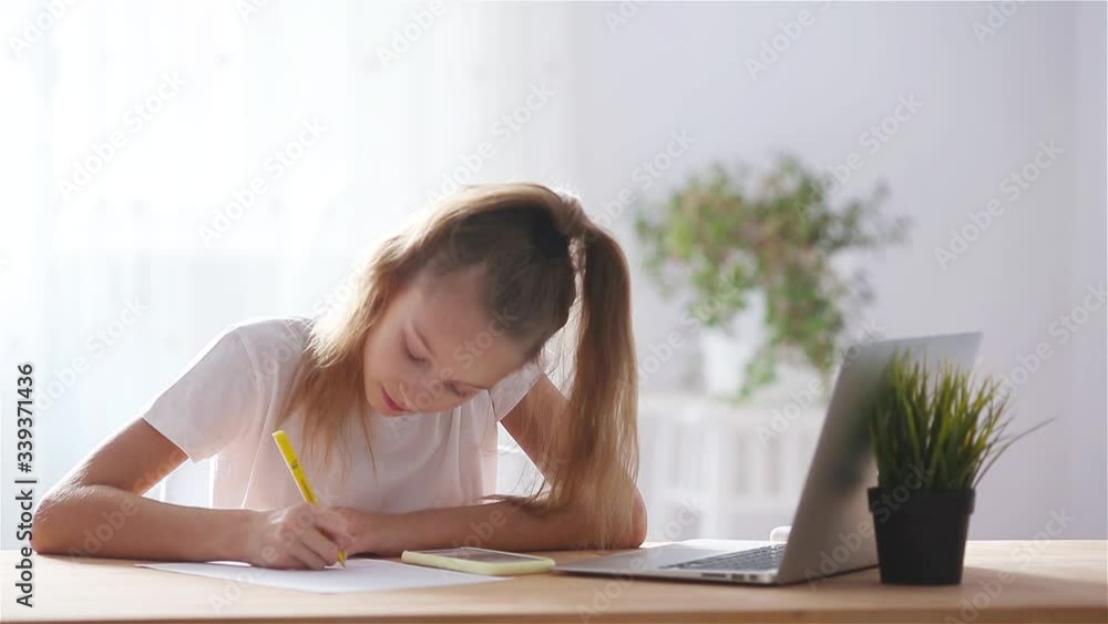 Serious schoolgirl sitting at table with laptop and textbook and doing homework. Serious schoolgirl sitting at table with laptop and textbook and doing homework.