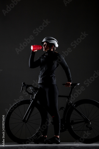 Girl posing on roadbike. Drinking from red cycling water bottle. White protective helmet. Side lit cyclist against dark background. © Nikola Spasenoski