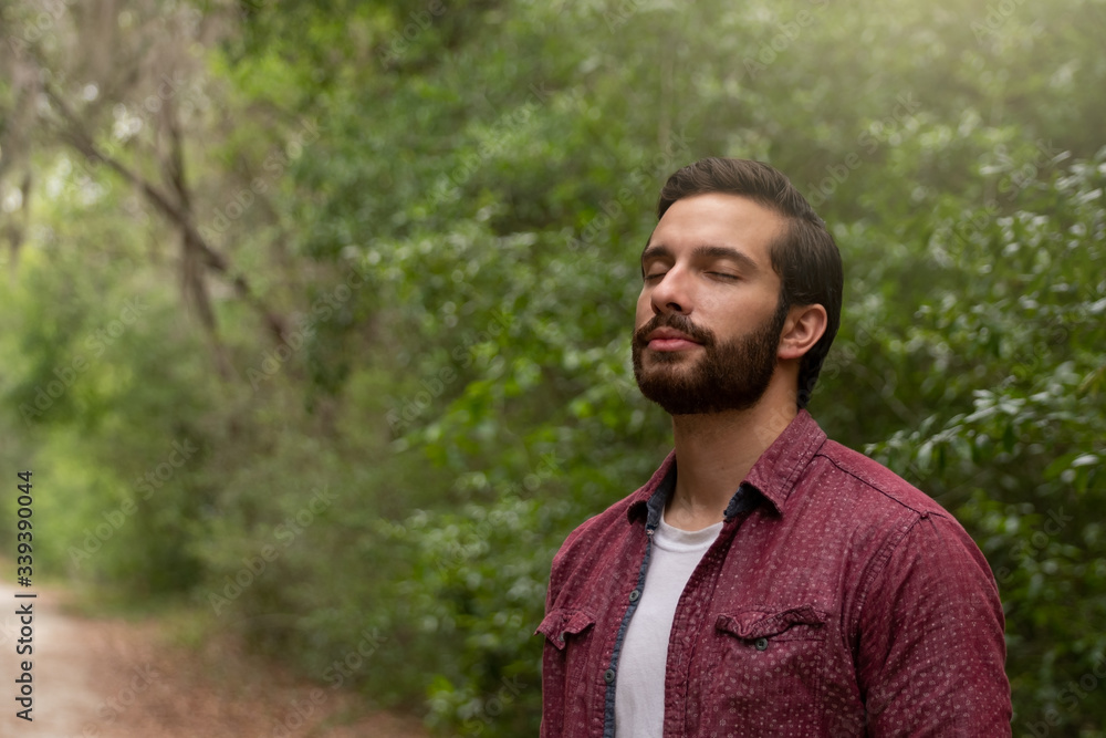 young man with brown hair and beard wearing red button down breathes in a breath of fresh air immersing himself in a nature path to social distance himself in order to stay healthy and alive. young man with brown hair and beard wearing red button down breathes in a breath of fresh air immersing himself in a nature path to social distance himself in order to stay healthy and alive.