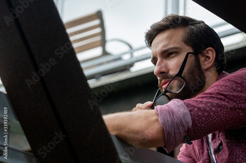 A bearded hipster man wearing a red button down shirt slightly taking off his glasses to see more clearly what is in the distance with a straight stare on his face squinting intensely outside.