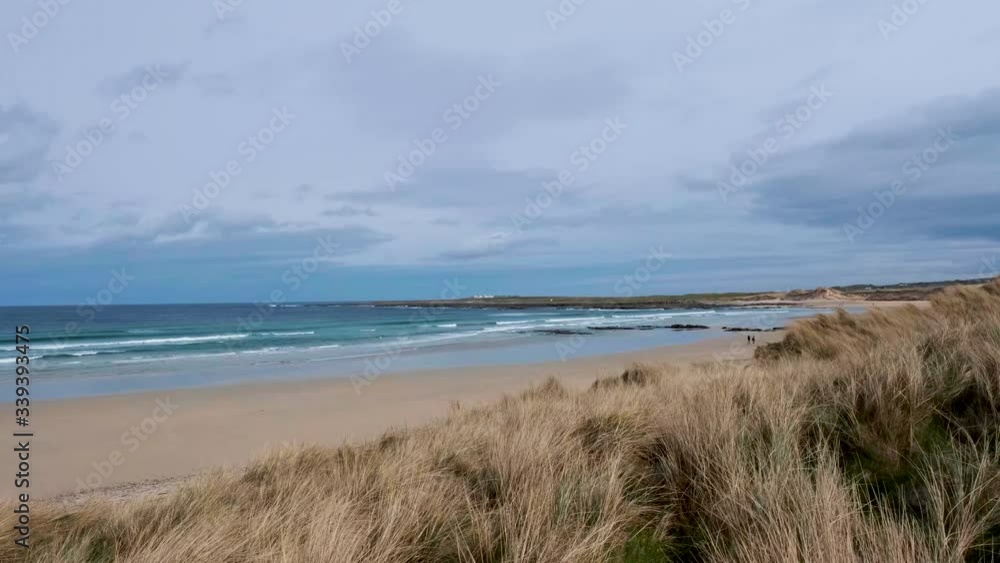 Couple in distance walks on beautiful lonely wide sandy beach behind grassy dunes on cloudy day waves rushing to shore. Panning shot slow motion Couple in distance walks on beautiful lonely wide sandy beach behind grassy dunes on cloudy day waves rushing to shore. Panning shot slow motion