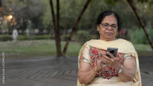 Senior Indian woman with glassses wearing traditional salwar kameez dialing and speaking on phone, sitting in a park in Delhi. Concept - Digital literacy for senior citizens of India photo