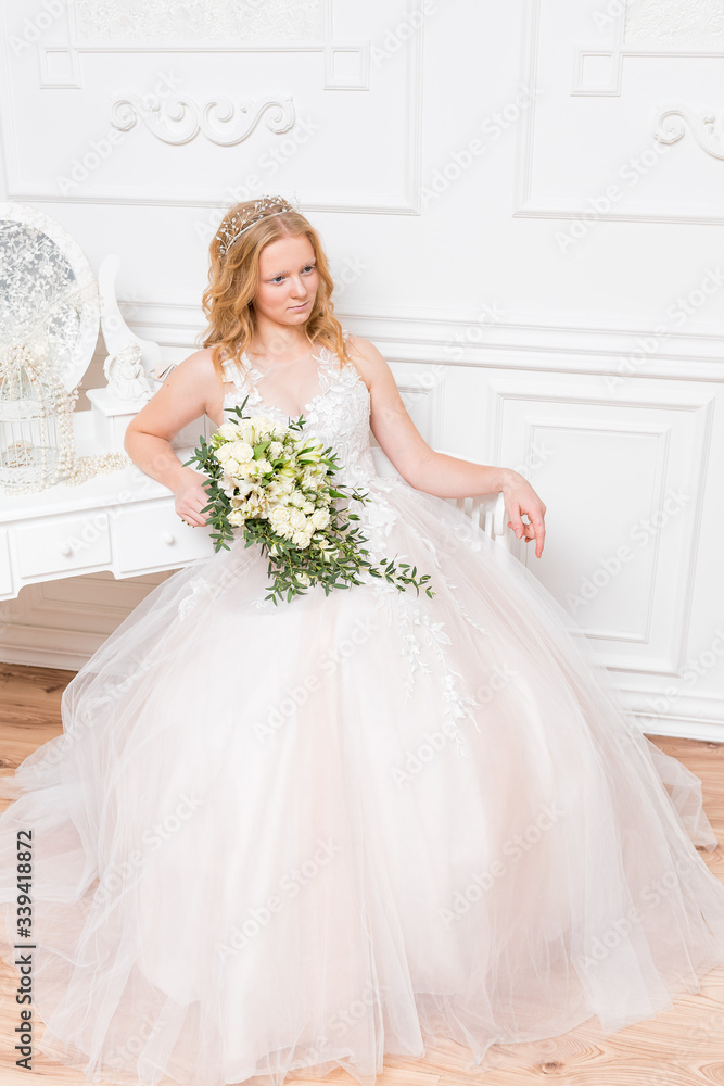 Romantic young bride in hotel room at elegant table in white lace wedding dress with makeup and wavy blonde hair with wedding bouquet in her hands getting ready in bride's morning. Fine art wedding. 