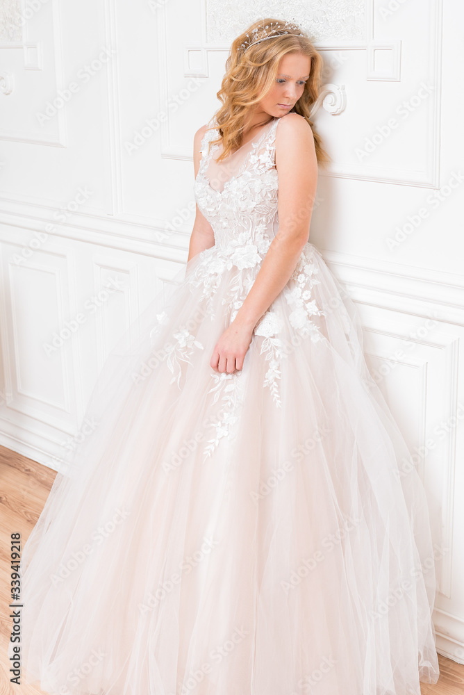 Romantic shy young bride in elegant hotel room in white lace wedding dress with makeup and wavy blonde hair with pearl jewelry getting ready in bride's morning. Fine art wedding. Romantic shy young bride in elegant hotel room in white lace wedding dress with makeup and wavy blonde hair with pearl jewelry getting ready in bride's morning. Fine art wedding.