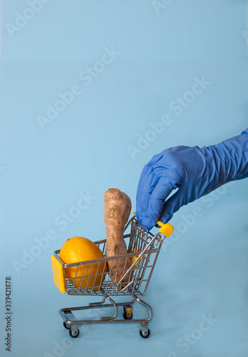 Female hand with a blue rubber glove rolls a supermarket cart with ginger and lemon on a blue background. Vitamins Health. Protection. covd 19 photo
