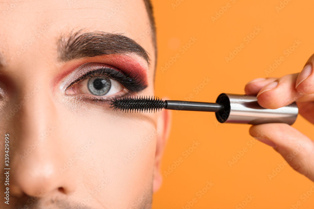 Young transgender woman applying mascara onto eyelashes against color background, closeup