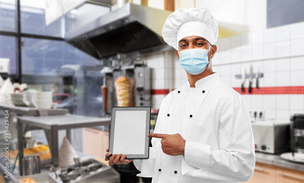 health, safety and pandemic concept - indian male chef cook in toque with tablet pc computer wearing face protective medical mask for protection from virus disease over restaurant kitchen background health, safety and pandemic concept - indian male chef cook in toque with tablet pc computer wearing face protective medical mask for protection from virus disease over restaurant kitchen background