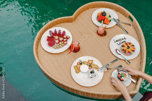 Breakfast tray in swimming pool, floating breakfast in luxury hotel. Girl relaxing in the pool drinking coffee and eating fruit plate by the hotel pool. Exotic summer diet. Tropical beach lifestyle