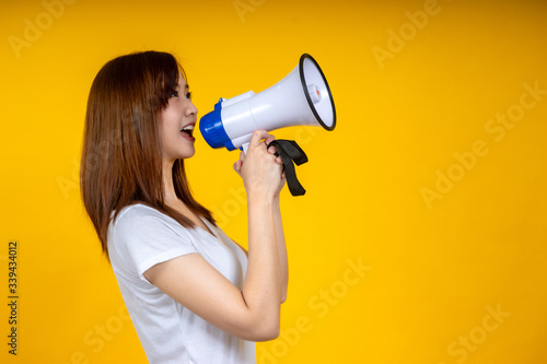 Young Asian woman in white casual t-shirt looking aside, scream in megaphone isolated on bright yellow wall background in studio. People lifestyle concept. Mock up copy space