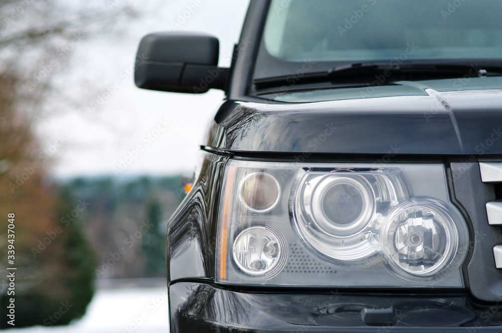 Modern luxury black premium SUV right car headlight with washer closeup over winter forest with snow background with copy space. Concept of expensive luxury premium sports offroad SUV. Modern luxury black premium SUV right car headlight with washer closeup over winter forest with snow background with copy space. Concept of expensive luxury premium sports offroad SUV.