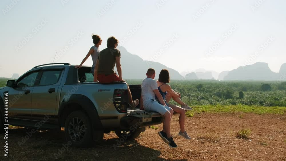 Group of tourists sitting in the back of a pickup truck and looking to the mountains during sunset. Friends inside a trunk car enjoying a road trip of mountains. Slow motion Group of tourists sitting in the back of a pickup truck and looking to the mountains during sunset. Friends inside a trunk car enjoying a road trip of mountains. Slow motion