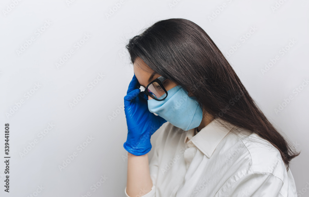 Young nurse doctor in a protective blue medical mask and gloves holding glasses in his hand and looking down. Physical fatigue, sleepiness. Concept of quarantine, pandemic, coronavirus COVID-19. Young nurse doctor in a protective blue medical mask and gloves holding glasses in his hand and looking down. Physical fatigue, sleepiness. Concept of quarantine, pandemic, coronavirus COVID-19.