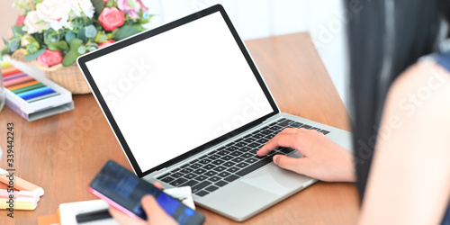Closeup image of stylish woman holding a smartphone and typing on computer laptop with white blank screen while sitting at the wooden working desk over comfortable living room as background.