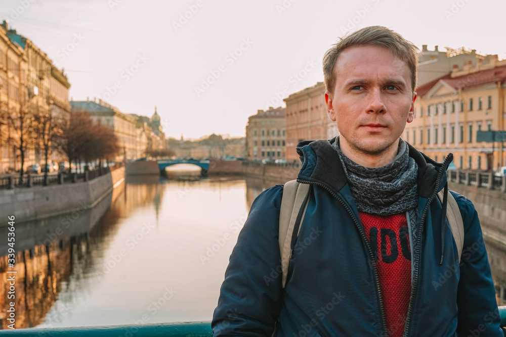 A man in a jacket and with a backpack stands on the Embankment of St. Petersburg, a beautiful dawn, a deserted city, empty streets, historical buildings attractions