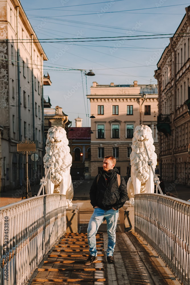 A young blond man in a jacket and with a backpack stands on the bridge with statues of white lions in the middle of the canals of St. Petersburg, Attractions, morning spring