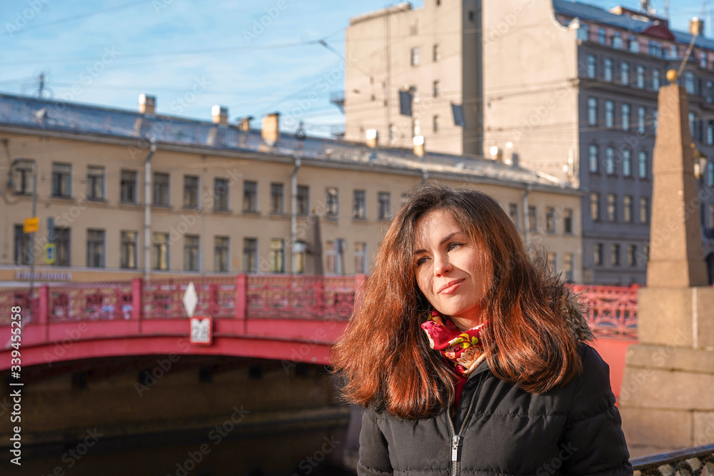 A girl with long hair in the middle of the street against the background of the Sights of St. Petersburg, a beautiful historical building near the red bridge near the river channel, morning spring