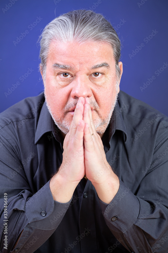 Man studio closeup portrait. He is serious and looking to the camera with his hands in front of his face. He looks preoccupied. The background is lighted with blue color. Man studio closeup portrait. He is serious and looking to the camera with his hands in front of his face. He looks preoccupied. The background is lighted with blue color.