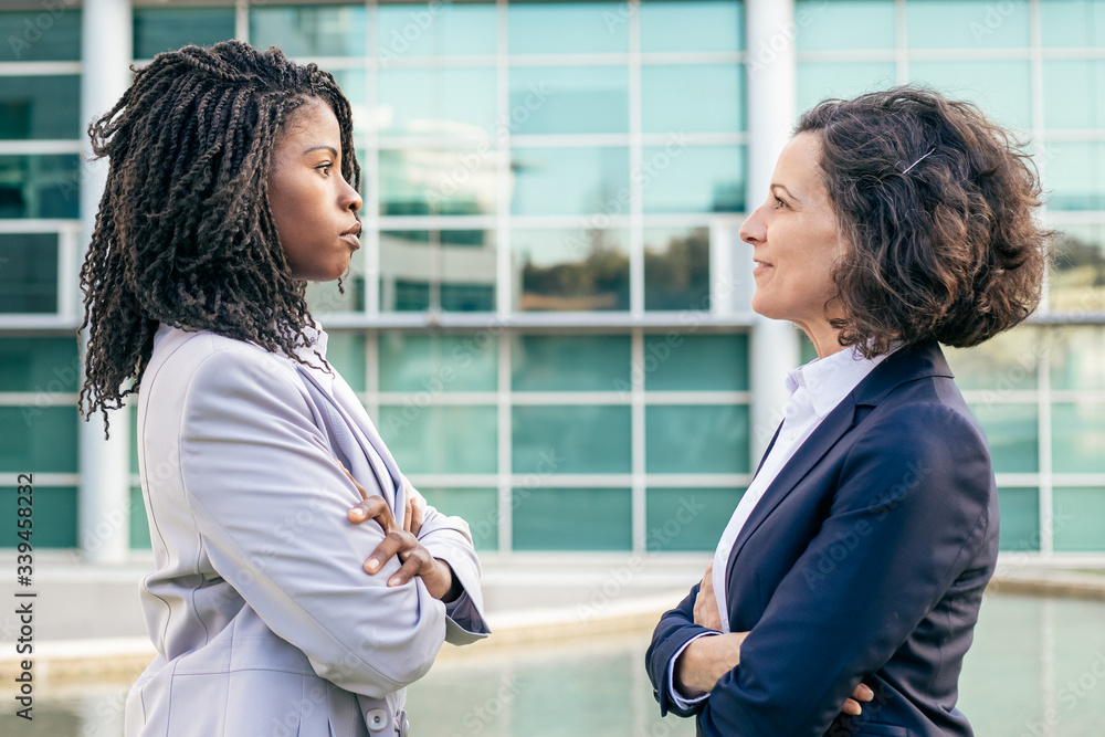 Coworkers standing with crossed arms and looking at each other. Side view of professional confident multiethnic businesswomen standing outside office building. Business concept Coworkers standing with crossed arms and looking at each other. Side view of professional confident multiethnic businesswomen standing outside office building. Business concept