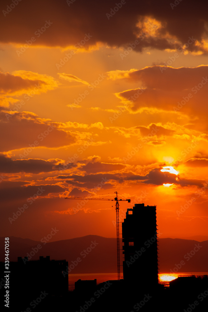 Construction crane at sunset on a background of orange sun, a city by the sea