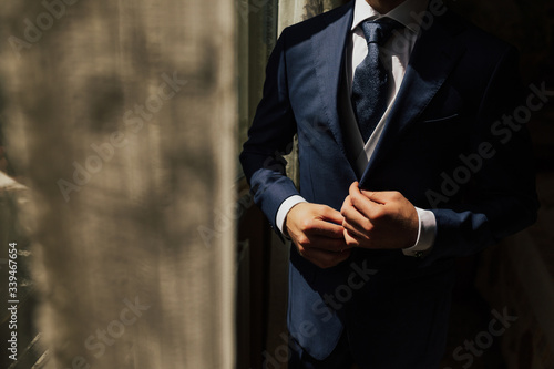 Grooms morning preparation. Cropped photo of elegant young fashion man dressing up for wedding celebration. Groom dressed in modern blue formal suit, white shirt getting ready for event. Sunny morning