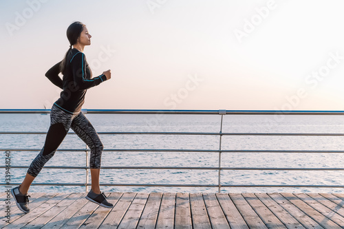 Athletic young woman jogging in the morning by sea wooden embankment. Silhouette of girl in sports costume. Beautiful sunlight. Healthy lifestyle concept. Copy space