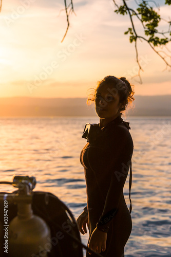 Female Scuba Diving Instructor Wearing a Wet Suit Standing Next to a Twin Tank at Sunset