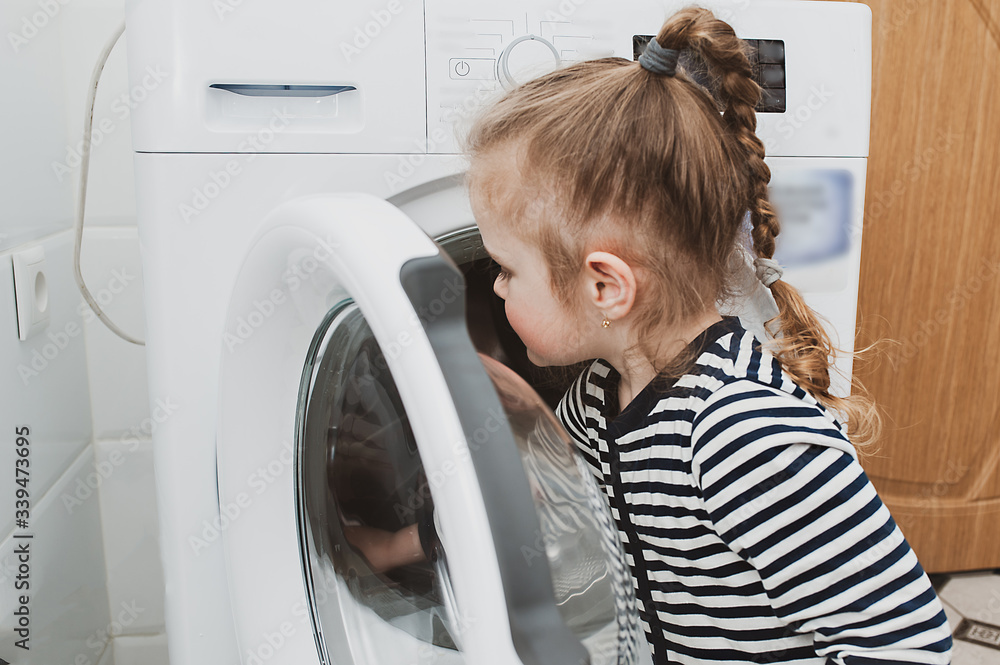 Cute girl in striped clothes puts things into the washing machine. Cleanliness and Household Concept Cute girl in striped clothes puts things into the washing machine. Cleanliness and Household Concept