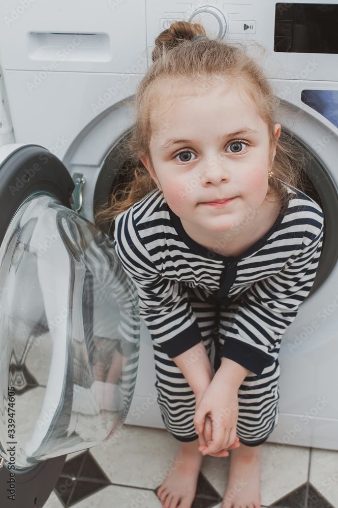 A cute girl in striped clothes sits inside a washing machine. Cleanliness and Household Concept A cute girl in striped clothes sits inside a washing machine. Cleanliness and Household Concept