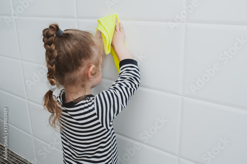 Cute girl in striped clothes washes a yellow rag on the wall. The child cleans at home. Household duties. Cute kid girl cleaning around. The idea of a child’s activity during quarantine