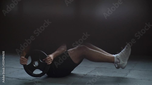 Athletic muscular man doing russian twist exercise with weight from barbell on black mat during sport workout training in modern dark gym. Concept of healthy lifestyle. Shooting in slow motion. photo