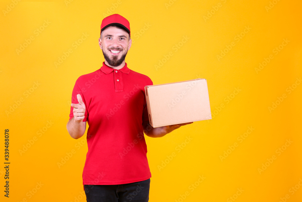 Young handsome delivery guy wearing red uniform and cap holding the blank cardboard box over isolated yellow background. Portrait of friendly bearded man carrying parcel. Copy space for text.