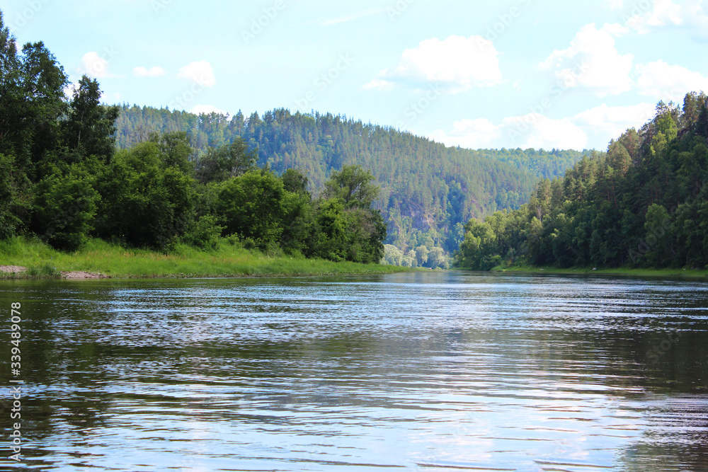 A river that flows through the forest. Mountain forest in the distance in a blue haze, river water landscape, sunny blue sky, Russia, South Ural, Belaya river, river rafting, travel.