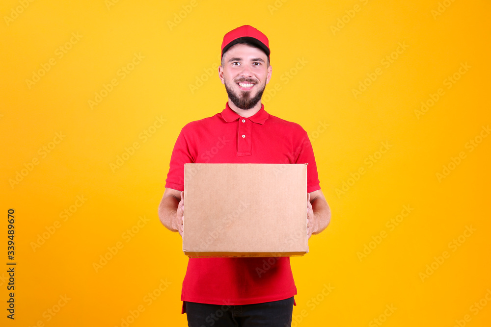 Young handsome delivery guy wearing red uniform and cap holding the blank cardboard box over isolated yellow background. Portrait of friendly bearded man carrying parcel. Copy space for text. Young handsome delivery guy wearing red uniform and cap holding the blank cardboard box over isolated yellow background. Portrait of friendly bearded man carrying parcel. Copy space for text.