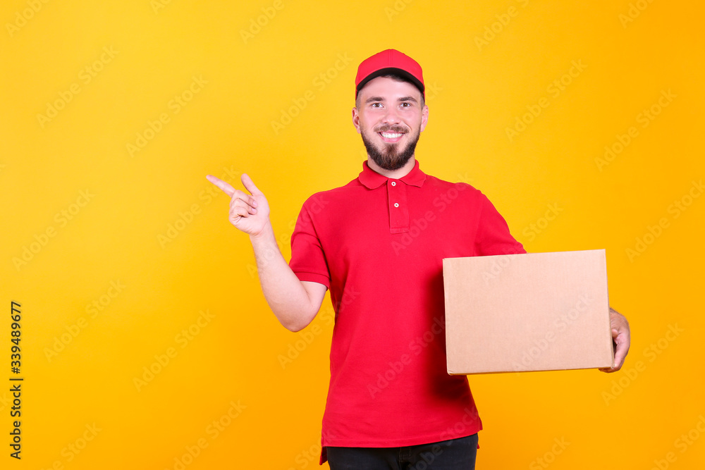 Young handsome delivery guy wearing red uniform and cap holding the blank cardboard box over isolated yellow background. Portrait of friendly bearded man carrying parcel. Copy space for text.