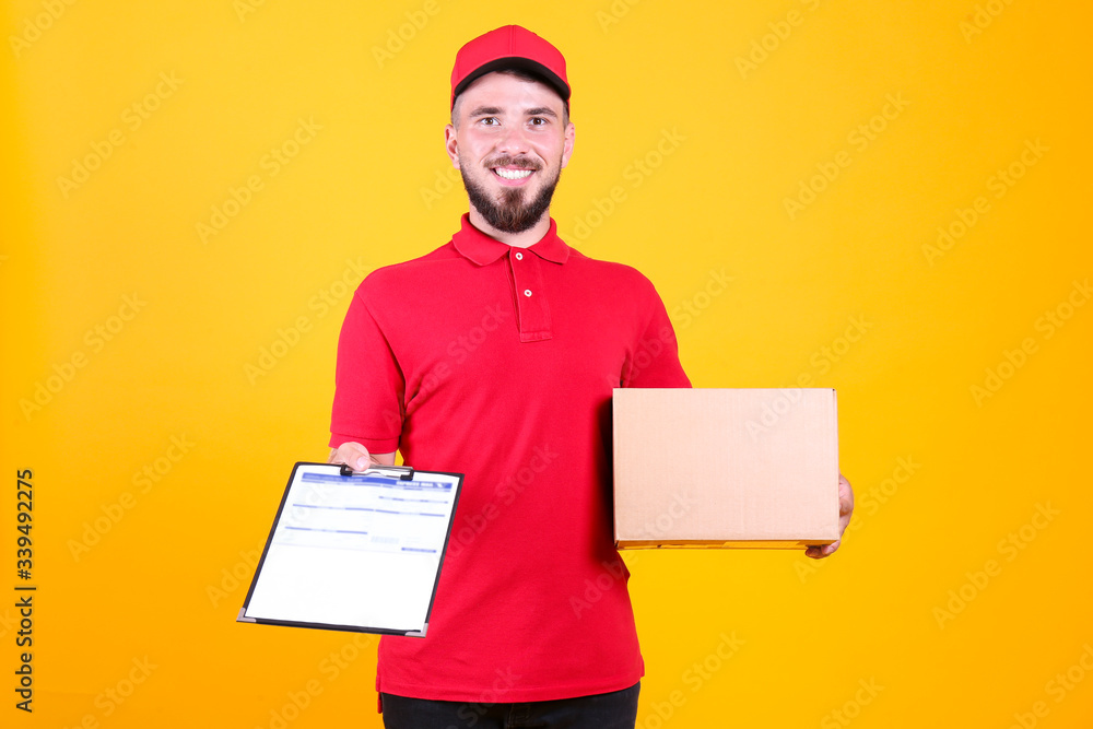 Young handsome delivery guy wearing red uniform and cap holding the blank cardboard box over isolated yellow background. Portrait of friendly bearded man carrying parcel. Copy space for text. Young handsome delivery guy wearing red uniform and cap holding the blank cardboard box over isolated yellow background. Portrait of friendly bearded man carrying parcel. Copy space for text.