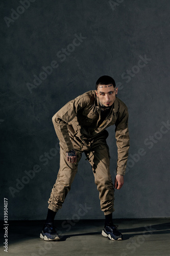 Young male with tattooed body and face, earrings, beard. Dressed in khaki jumpsuit and black sneakers. Dancing on gray background. Dancehall, hip-hop