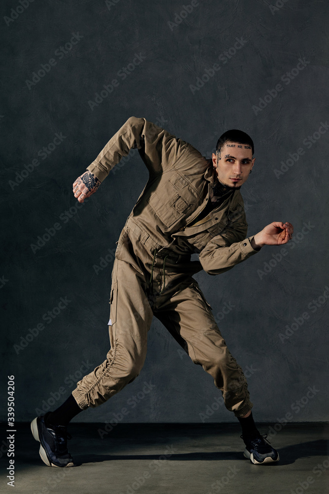 Young fellow with tattooed body and face, earrings, beard. Dressed in khaki jumpsuit, black sneakers. Dancing on gray background. Dancehall, hip-hop