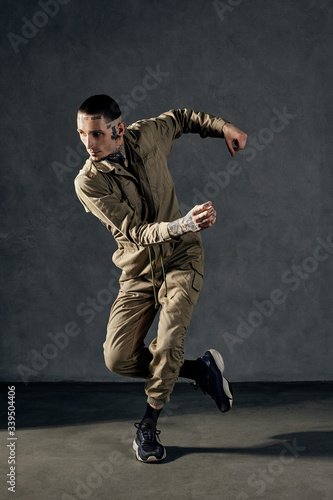 Modern guy with tattooed body and face, earrings, beard. Dressed in khaki overalls and black sneakers. Dancing on gray background. Dancehall, hip-hop