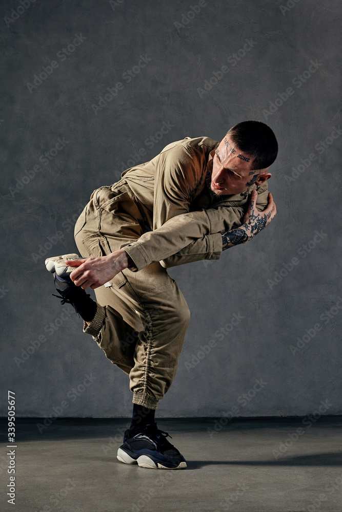 Attractive man with tattooed body and face, earrings, beard. Dressed in khaki overalls, black sneakers. Dancing on gray background. Dancehall, hip-hop