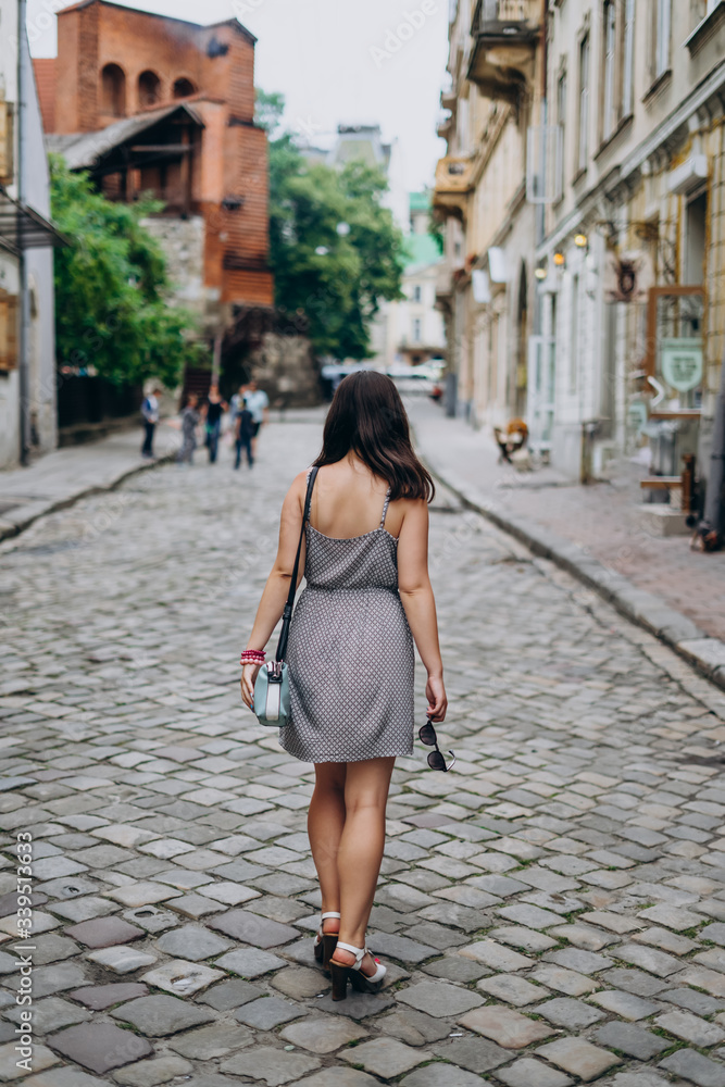Young woman in a gray sundress on a city tour. Brunette woman in dress and sandals walking on city streets. Girl in a handbag and glasses.