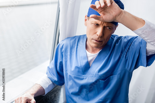 Tableau sur toile tired and middle aged doctor in scrub hat touching forehead near window in hospi
