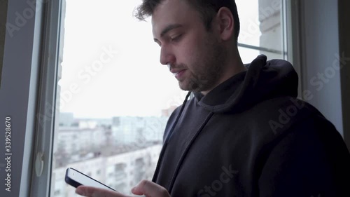 Amazed young man checks his phone, smoking iqos by the window, close up