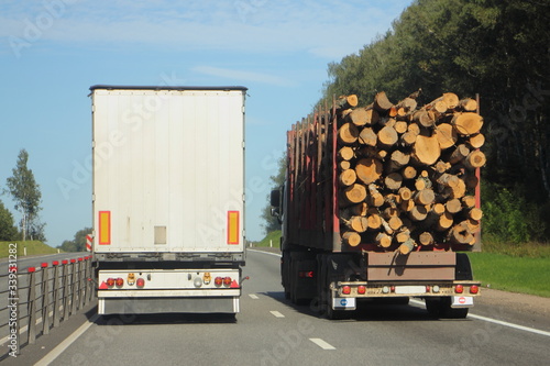 Semi truck with white van trailer overtakes loaded timber truck on suburban highway, rear view from road at summer day on clear blue sky and forest background, cargo logistics