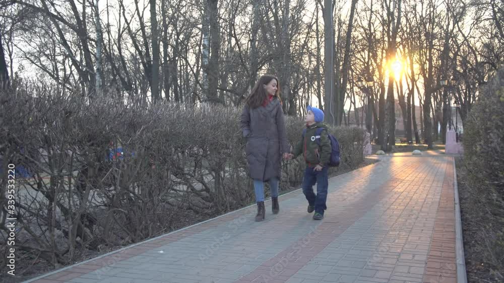 Wide shot of young Caucasian woman and schoolboy walking along the alley in park on sunset and talking. Portrait of happy mother and son on evening stroll leaving. Lifestyle, joy, unity. Wide shot of young Caucasian woman and schoolboy walking along the alley in park on sunset and talking. Portrait of happy mother and son on evening stroll leaving. Lifestyle, joy, unity.